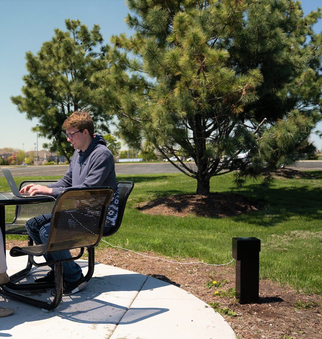A person sitting at an outdoor table using a laptop while it's being charged with Pedoc power pedestal